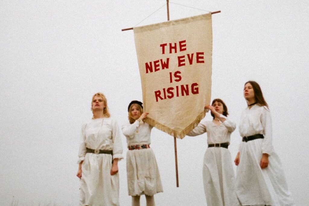 Particolare della copertina dell'abum The New Eve is Rising della band The New Eves. Le quattro componenti del gruppo sono vestite di bianco su sfondo bianco e tengono uno stendardo bianco su cui è scritto il titolo dell'album in rosso