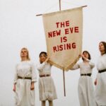 Particolare della copertina dell'abum The New Eve is Rising della band The New Eves. Le quattro componenti del gruppo sono vestite di bianco su sfondo bianco e tengono uno stendardo bianco su cui è scritto il titolo dell'album in rosso