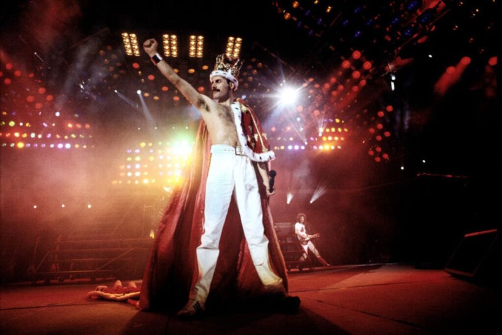 Freddy Mercury in un live, con il suo costume regale in tipico stile glam rock e il resto della band sullo sfondo
