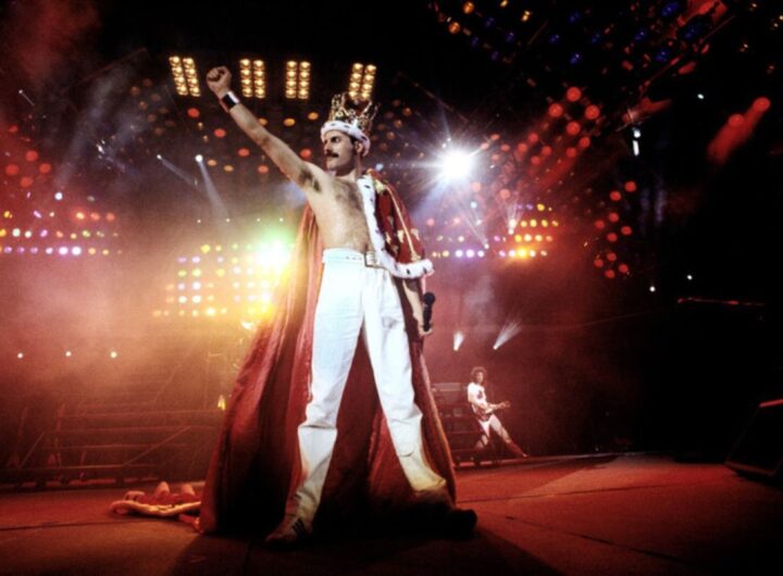 Freddy Mercury in un live, con il suo costume regale in tipico stile glam rock e il resto della band sullo sfondo